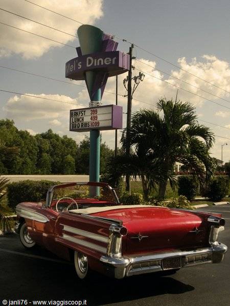 Classic red convertible parked under Mel's Diner sign among palm trees