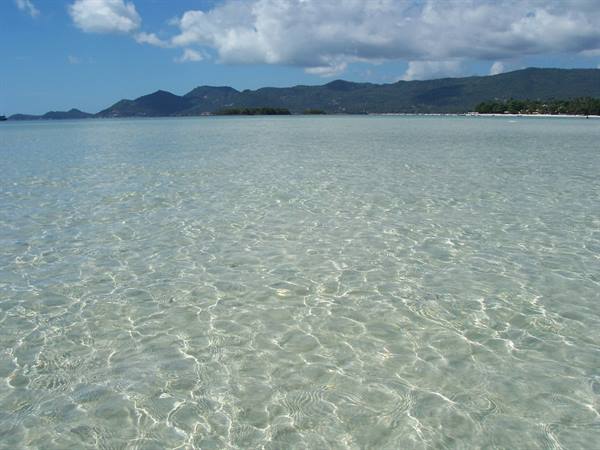Shallow crystal-clear water over light sand, green hills along the coast