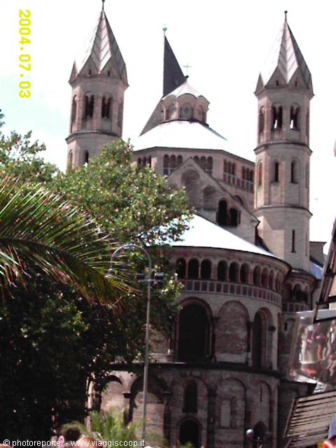 Romanesque stone cathedral with two pointed spired towers and semicircular apse, trees in front