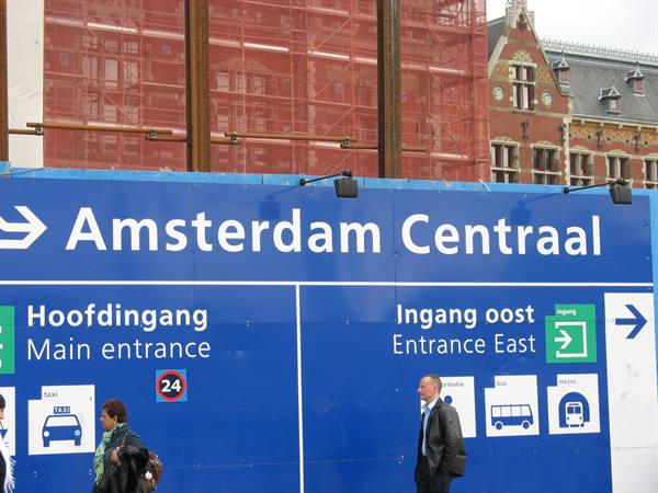 Blue Amsterdam Centraal sign with pedestrians, red scaffolding and old station behind