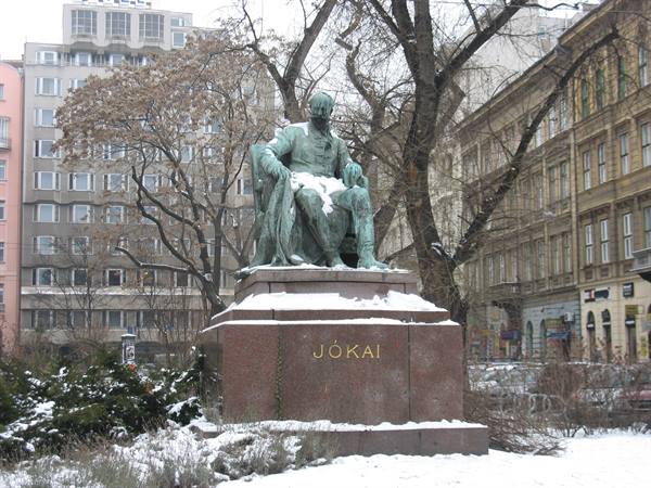 Bronze statue of Jókai on snowy granite pedestal among city buildings