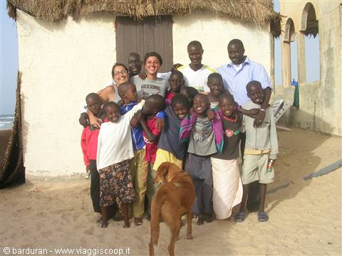 Group of adults and children smiling in front of thatched hut on beach, dog nearby