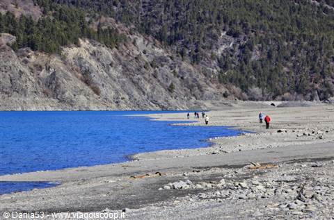 Walkers on a gravel lakeshore, blue water below forested rocky slopes