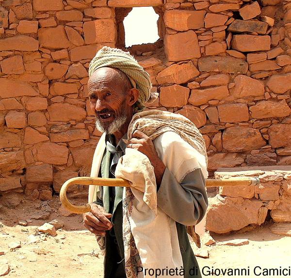 Elderly man with turban and staff before red stone wall