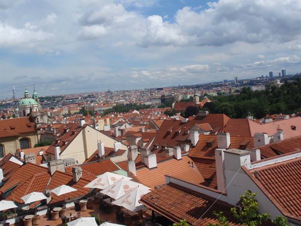 Red-tiled rooftops over Prague, terrace umbrellas below, white clouds above