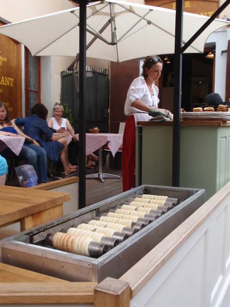 Woman baking chimney cakes on rotating rollers, customers seated under the umbrella