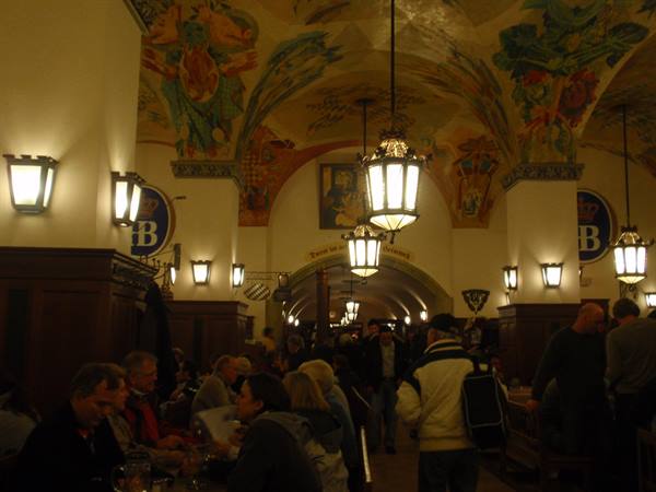 Painted hall of Munich's Hofbräuhaus, lit lanterns, people seated at wooden tables