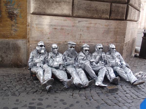Six street performers painted silver sitting beside a travertine wall