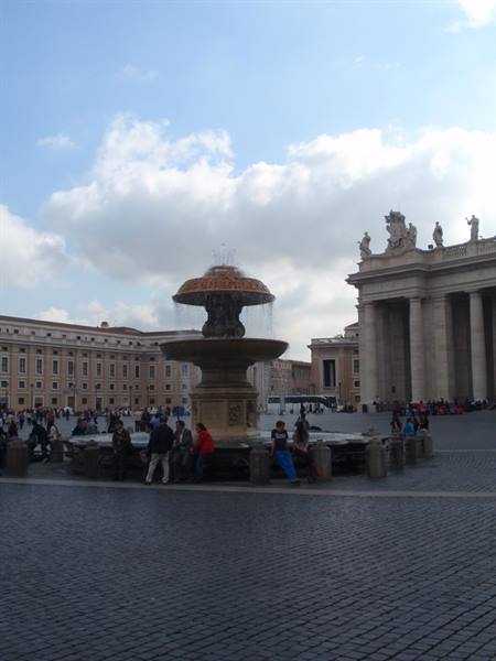 Fountain in St. Peter's Square, tourists sitting around under cloudy sky