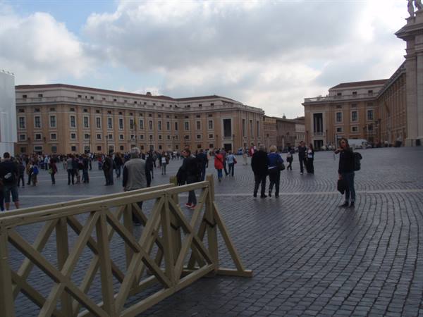 Tourists on cobblestones in St. Peter's Square, Vatican building, wooden barrier, cloudy sky