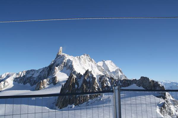 Snow-covered alpine peaks under blue sky, metal fence in foreground, Chamonix