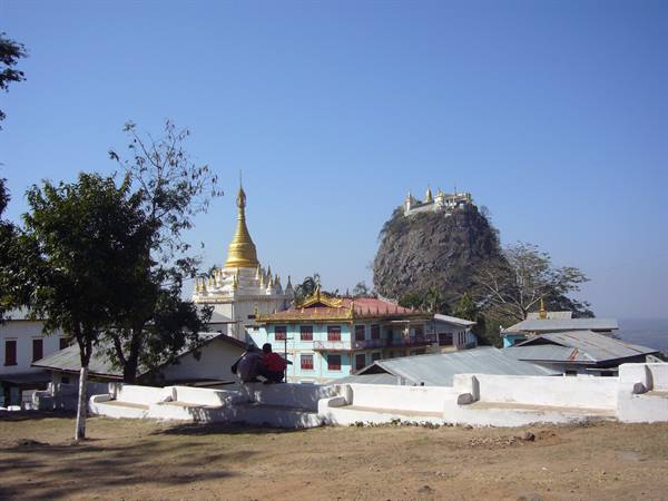 Golden stupa and monastery below Mount Popa's rock, two people sitting nearby