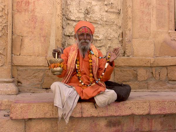 Orange-robed sadhu blessing with a brass pot before Jaisalmer's sandstone wall