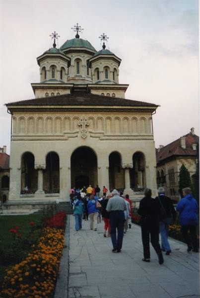 Tourists walk toward Alba Iulia cathedral among flowerbeds at dusk