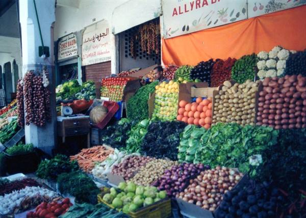 Market stall piled with fruit and vegetables, Arabic signs, vendor in shade
