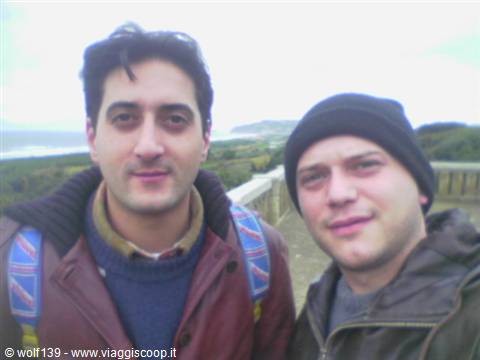 Two backpackers smile in front of a stone balustrade above the coastline