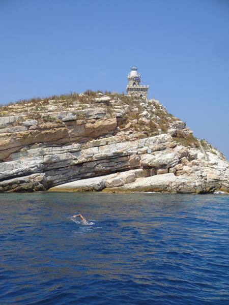 Swimmer in blue sea below rocky islet with stone lighthouse