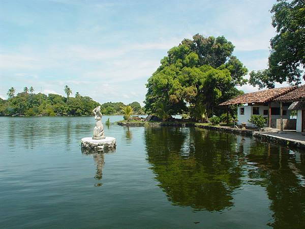 White statue on tiny islet, tiled-roof house and lush trees by the lake