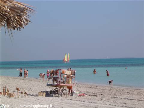 White-sand beach: vendor cart with fabrics, swimmers and yellow-sailed catamaran in turquoise water