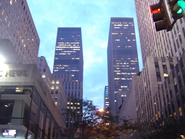 Rockefeller Center skyscrapers at dusk, lit windows, traffic light in foreground
