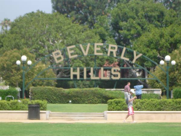 Beverly Hills metal sign among trees, people walking across the sunny lawn