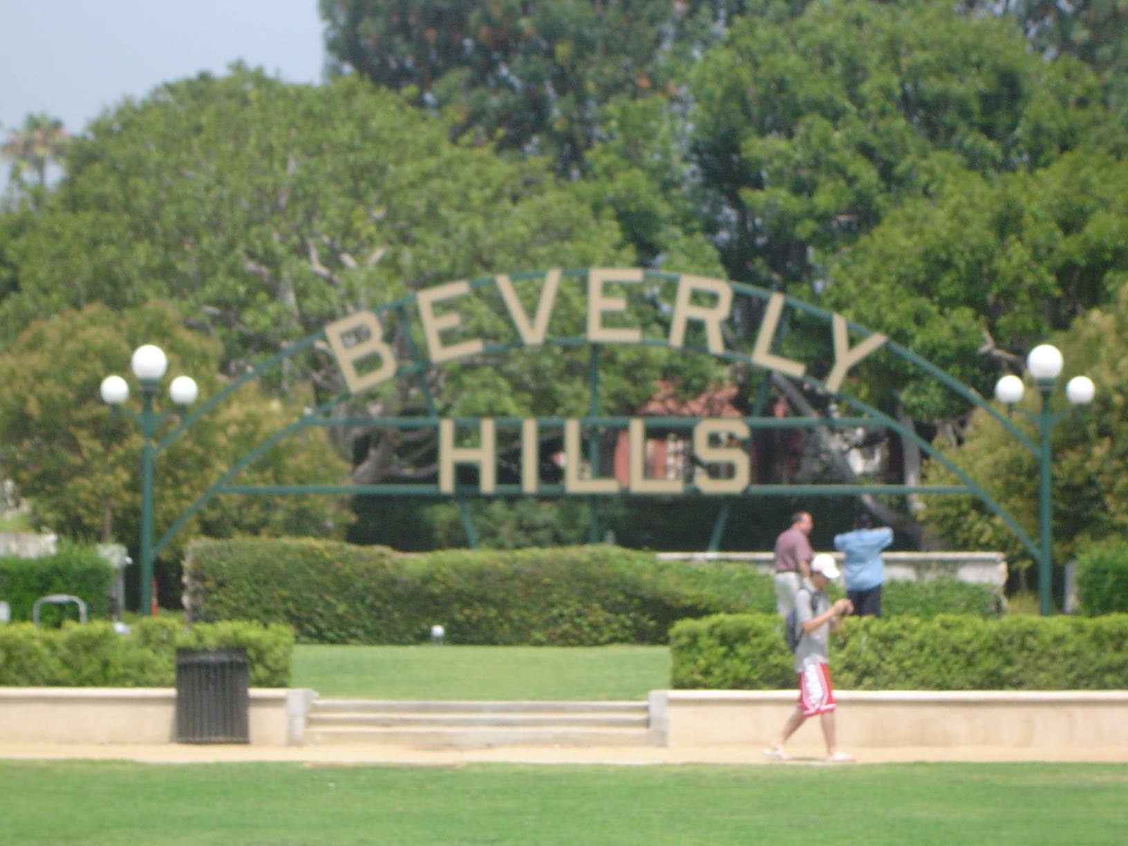 Beverly Hills metal sign among trees, people walking across the sunny lawn