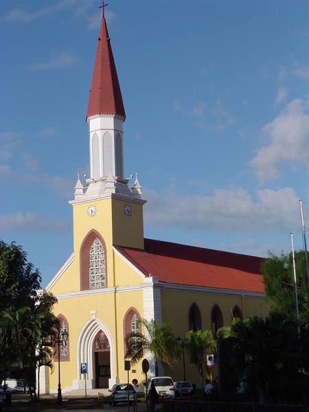 Yellow cathedral with clock tower and red spire, palm trees, cars, blue sky