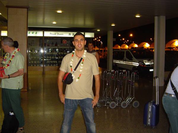 Traveler wearing flower lei at airport arrival, luggage trolleys and police van behind