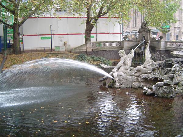 Stone statue with trident spouts a water jet in the fountain among trees