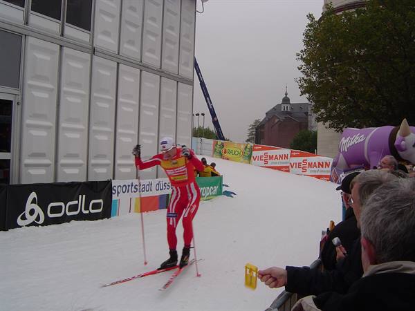 Cross-country skier in red suit beside white containers, fans with inflatable Milka cow