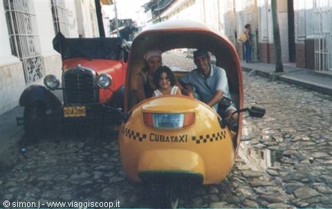 Smiling tourists riding a yellow Cuban cocotaxi on cobblestone street, vintage red car nearby