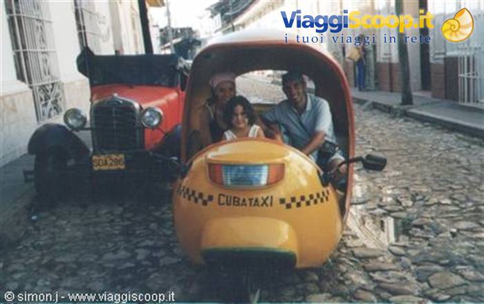 Smiling tourists riding a yellow Cuban cocotaxi on cobblestone street, vintage red car nearby