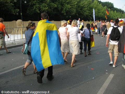 Man wearing Swedish flag walking among crowd on tree-lined street