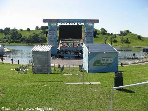 Stage with big screen beside the lake, white and blue structures on green grass
