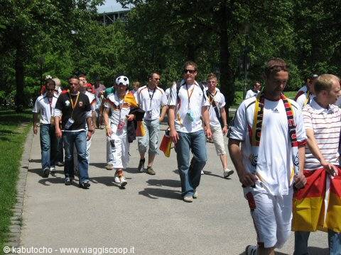 German football fans in black, red and gold scarves walk along a tree-lined path