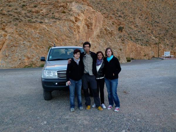 Four people beside a gray SUV on gravel, ochre rocks behind