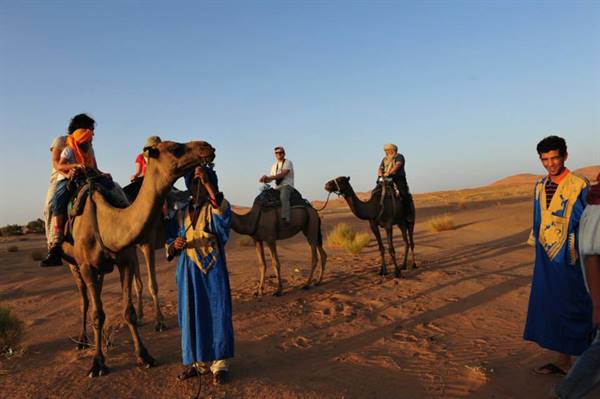 Tourists riding camels in desert, guide in blue robes nearby
