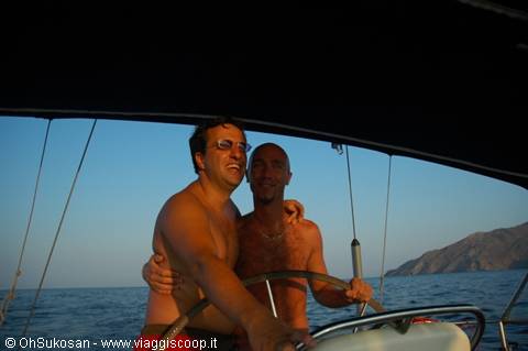 Two friends at the helm of a sailboat, sea and rocky coast behind