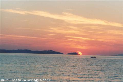 Red sun sinking into calm sea, dark islets and a small fishing boat