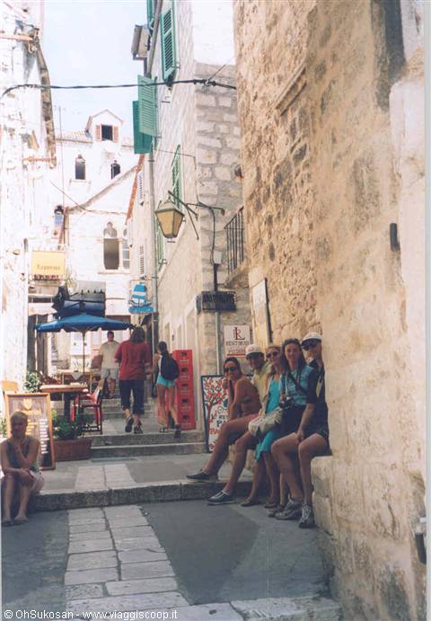 Tourists sitting on a stone wall in an alley with steps and green shutters
