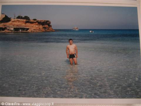 Man in black swimwear standing in clear shallow water, rocky coast behind