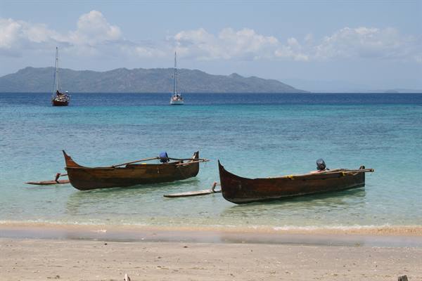 Two wooden outrigger canoes with outboard motors on the beach, sailboats and island beyond