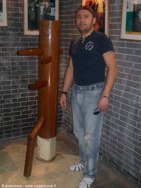 Man standing next to a wooden Wing Chun dummy against gray brick wall