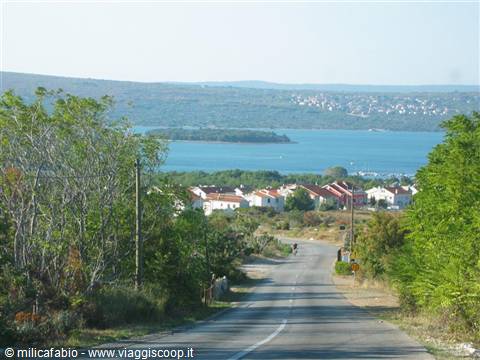 Asphalt road descends toward red-roofed houses and turquoise sea among green trees