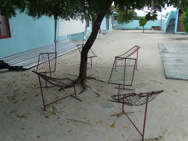 Empty metal framed mesh chairs under a tree, sand and blue houses