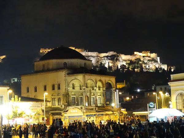 Monastiraki square at night, crowded, with the illuminated Acropolis behind