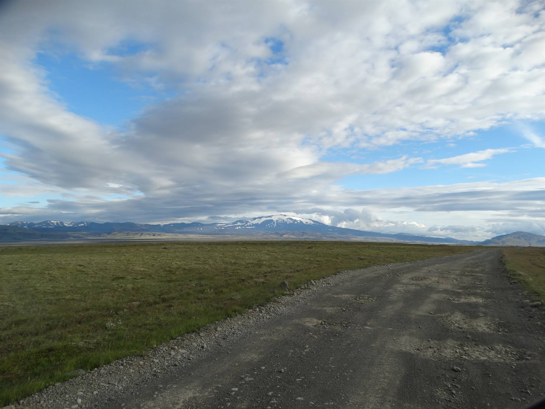 Strada sterrata tra praterie verdi verso montagne innevate sotto nuvole