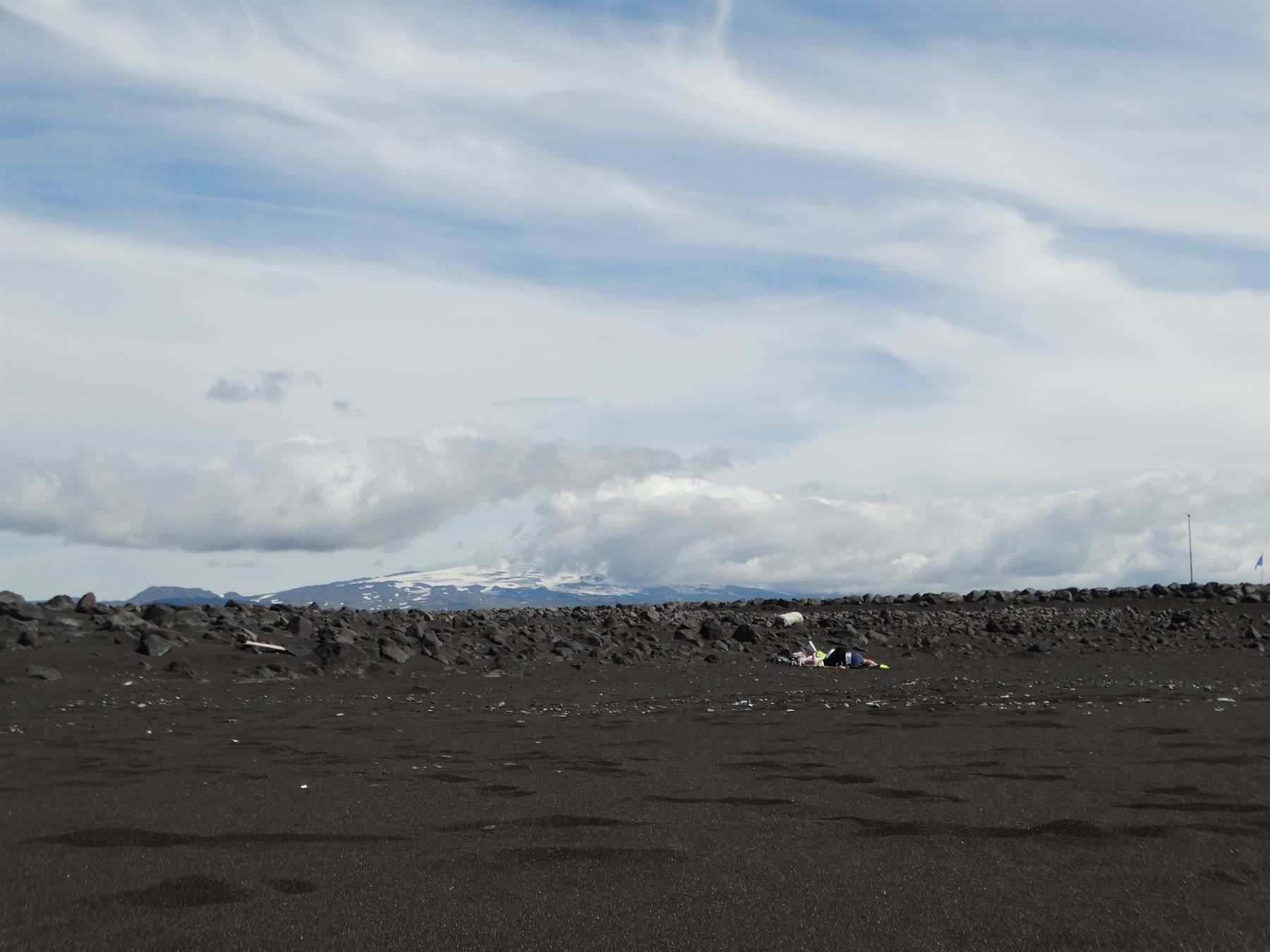 Spiaggia di sabbia nera vulcanica, rocce scure e vulcano innevato sotto nuvole