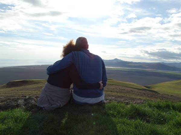 Couple sitting embraced on green grass, valley and mountains under cloudy sky