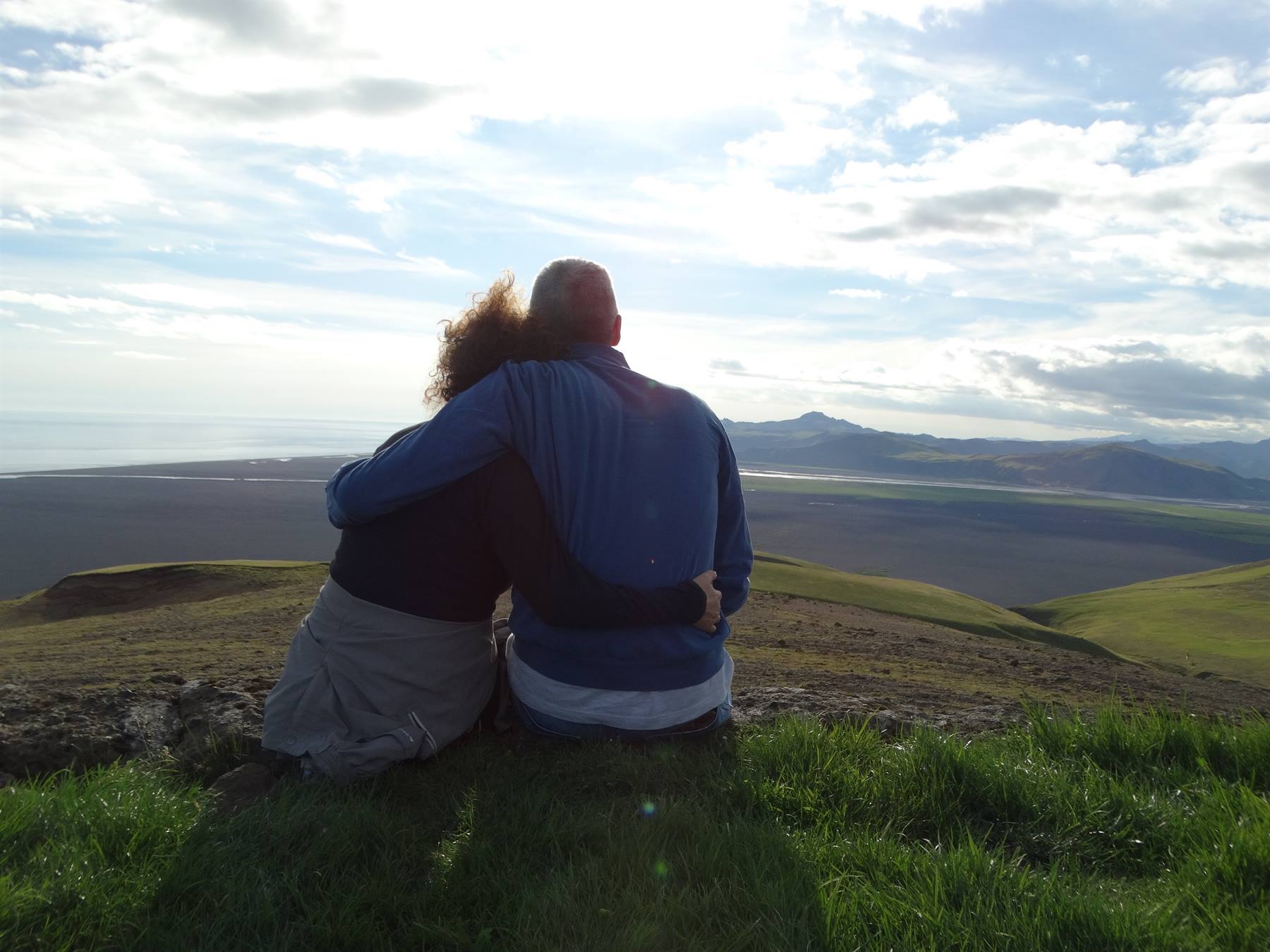 Couple sitting embraced on green grass, valley and mountains under cloudy sky